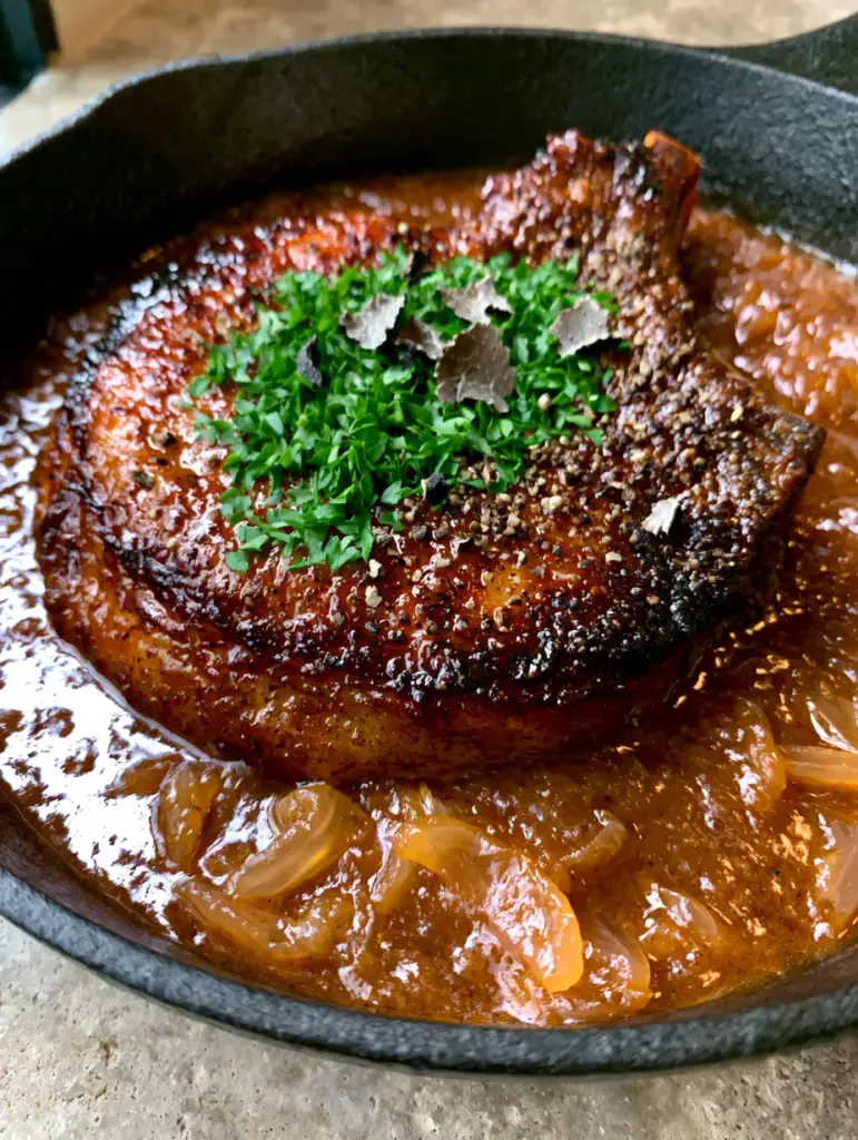 Smothered Pork Chops Recipe with Mahogany Onion Gravy 1 Thick bone-in pork chop in a cast iron skillet garnished with parsley and cracked black pepper. (smothered pork chops recipe)
