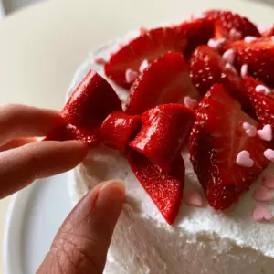 Extreme close-up of a hand placing a glossy red fondant bow onto a white cake with pink sprinkles. (Hello Kitty Birthday Party Ideas)