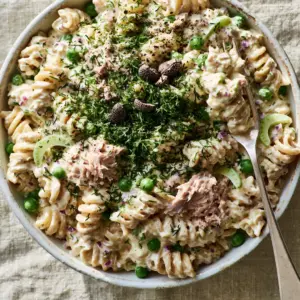 Macro shot of rotini spirals coated in a glossy ivory dressing with chunky tuna flakes and garden peas. (Tuna pasta salad)