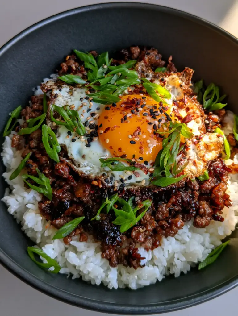 Korean Beef Bowl with Crispy Fried Eggs and Glossy Mahogany Glaze 1 Close up of steaming white jasmine rice topped with mahogany-colored Korean beef, scallions, and sesame seeds.