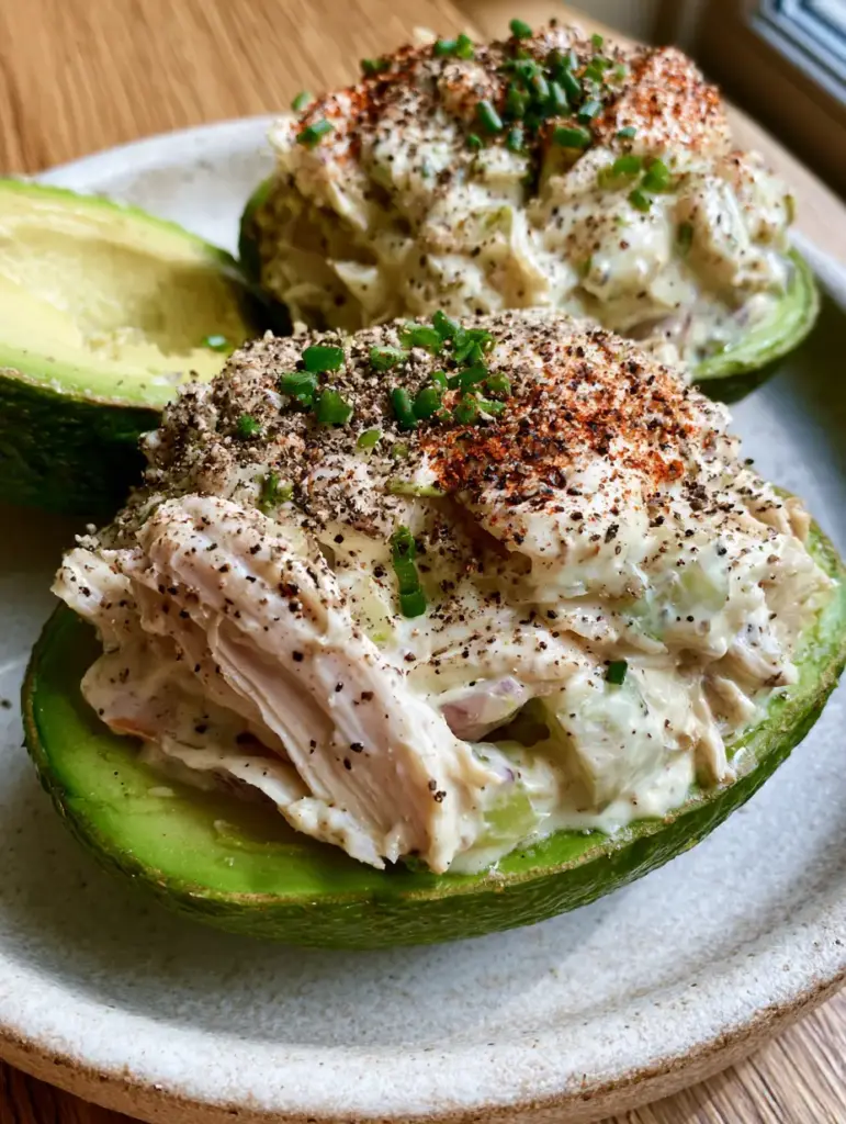 Chicken salad stuffed avocado with Creamy Greek Yogurt Dressing 1 Top-down view of Chicken salad stuffed avocado garnished with cracked black pepper and fresh microgreens on a wood surface.