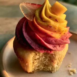 Close-up eye-level shot of a mango strawberry sunset cupcake showing the moist pale gold crumb and tall velvety ombre swirl with visible strawberry seeds.