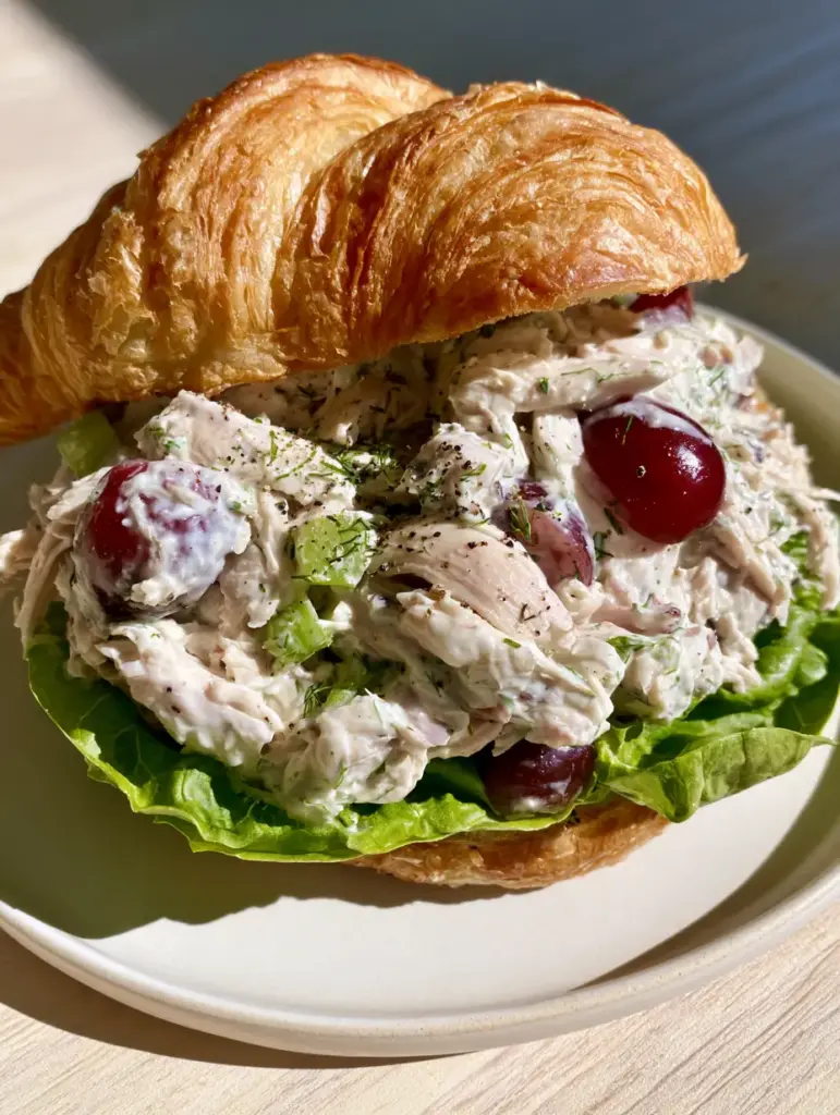 Chicken Salad Sandwich on Flaky Toasted Buttery Croissants 1 A Chicken Salad Sandwich on an off-white plate, highlighting the juicy red grapes and fresh bibb lettuce.