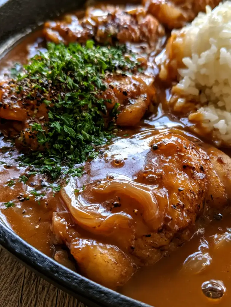 Southern Smothered Chicken with Velvety Brown Gravy and Caramelized Onions 2 Macro shot of golden-brown chicken skin partially submerged in a glossy, deep-brown gravy with caramelized onions. (Southern Smothered Chicken)