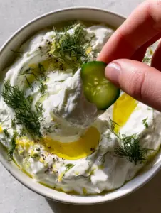 High-angle POV shot of a minimalist white bowl filled with velvety Greek yogurt cucumber dip garnished with dill and lemon zest.
