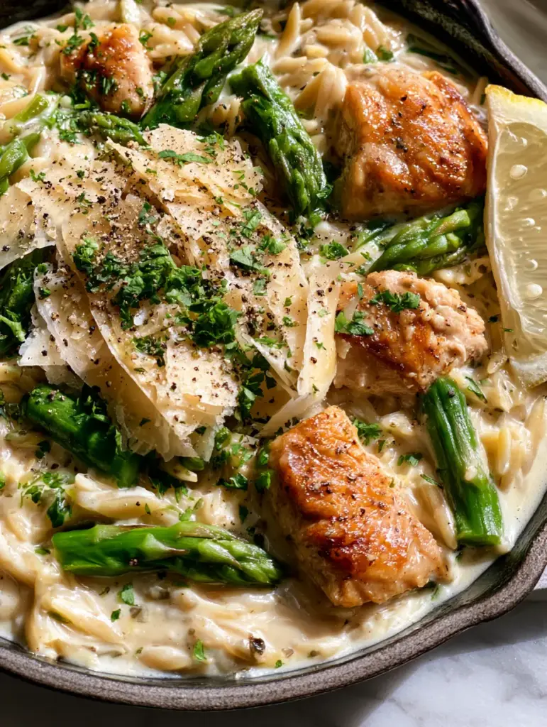 Close up of creamy parmesan orzo with chicken and asparagus garnished with coarse black pepper and large parmesan cheese shavings.