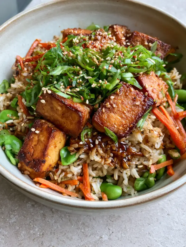 Tofu Fried Rice with Crispy Golden Cubes and Umami Glaze 1 Top-down view of a ceramic bowl filled with tofu fried rice garnished with sesame seeds and green scallions.