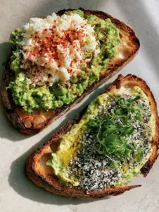 Top-down view of two slices of charred sourdough breakfast avocado toast on a stone countertop. (breakfast avocado toast)