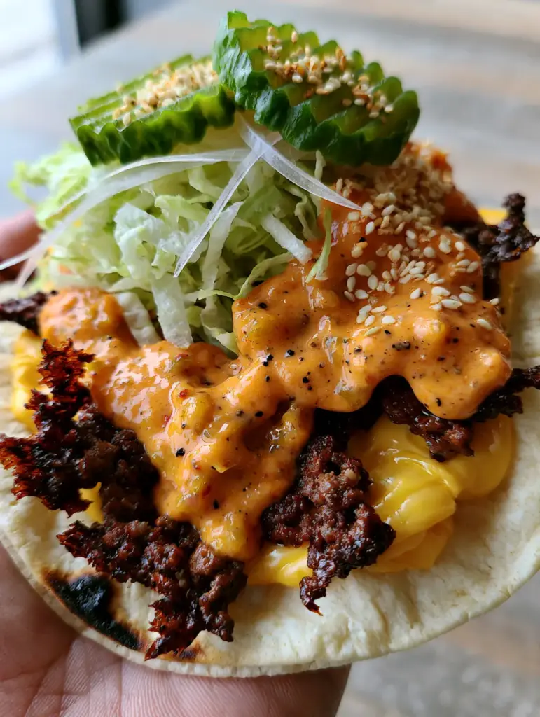 Smash Tacos: The Ultimate Guide to Crispy, Caramelized Tortilla Burgers 1 A handheld Smash Taco topped with creamy orange sauce, shredded iceberg lettuce, white onions, and crinkle-cut pickles.