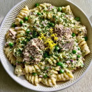Close up of al dente fusilli coated in thick glossy ivory dressing with moist tuna flakes and bright green peas.