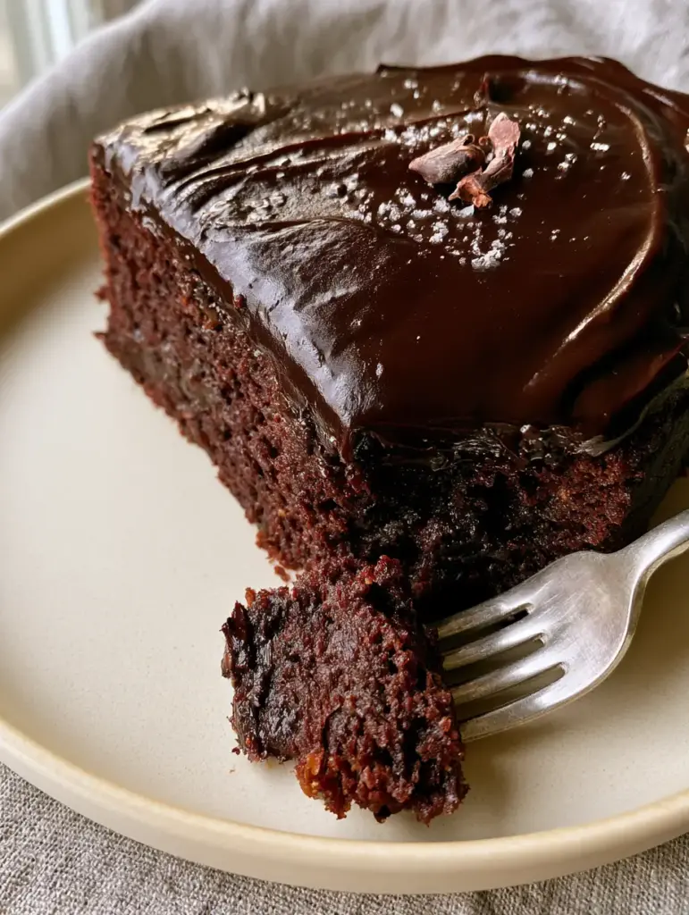 Vegan Chocolate Cake Recipe: The Ultimate Fudgy Dark Chocolate Masterpiece 1 A silver fork pressing into a slice of dark chocolate cake with wavy ganache and sea salt garnish.