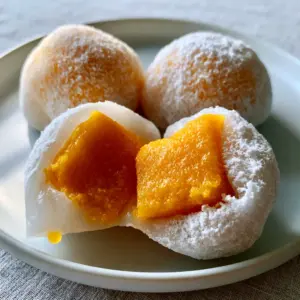 Extreme macro close-up of three plump mango mochi balls with a fine dusting of cornstarch on a white plate. (mango mochi)