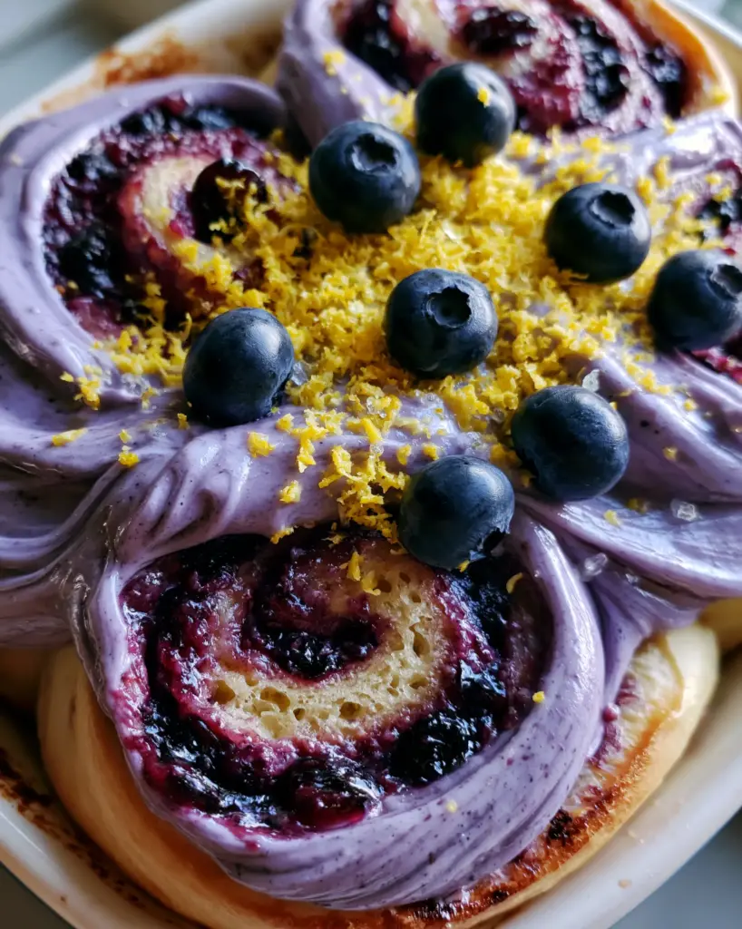 Blueberry Cinnamon Rolls with Lavender Cream Cheese Frosting 1 Close-up of pale lavender cream cheese frosting swirls on golden-brown sweet rolls with lemon zest and sugar crystals. (blueberry cinnamon rolls)