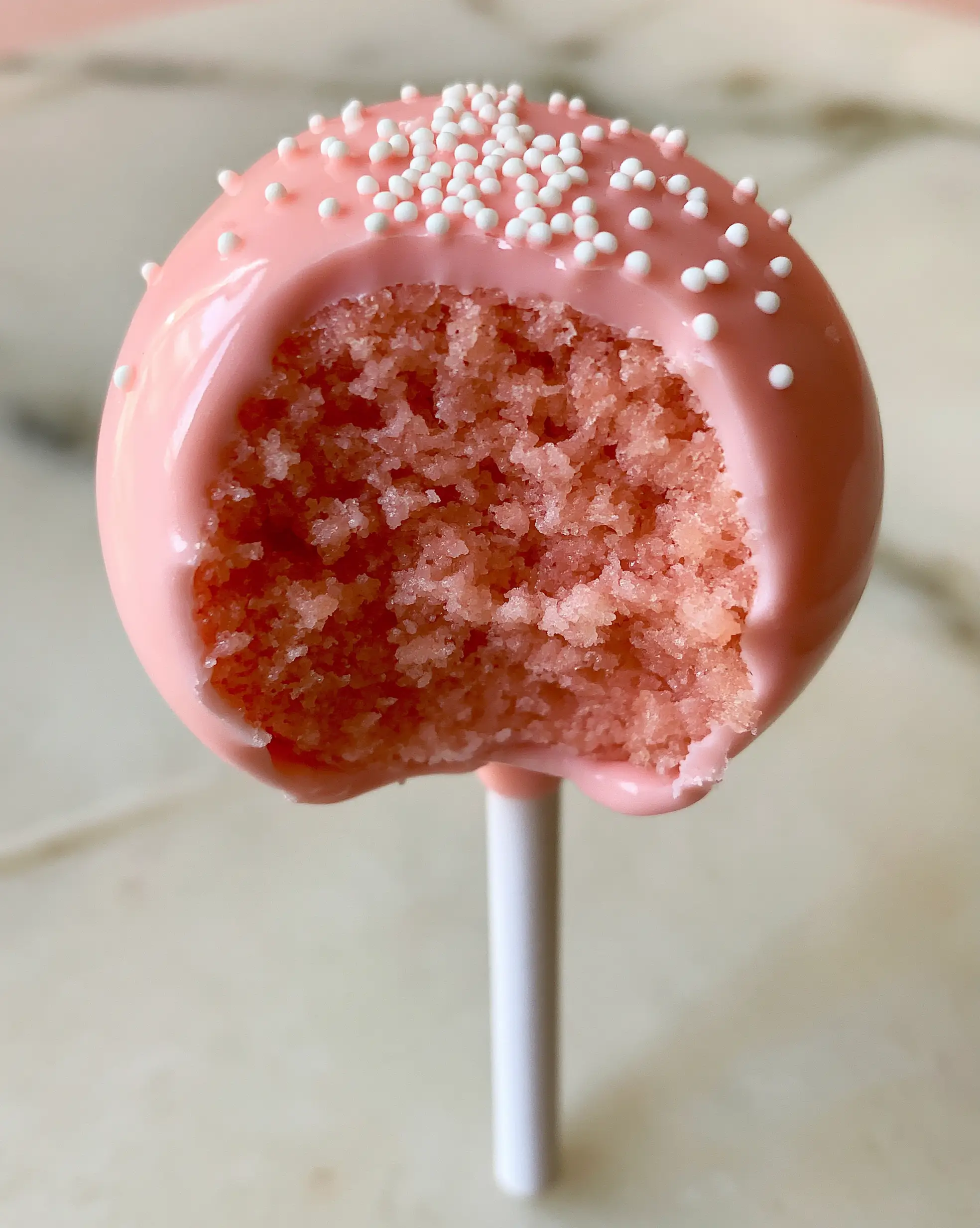 Cake Pop Flavors: The Ultimate Starbucks Copycat Pink Vanilla Recipe 3 A handheld smartphone photo of a pink cake pop with white nonpareils on a marble background. (cake pop flavors)